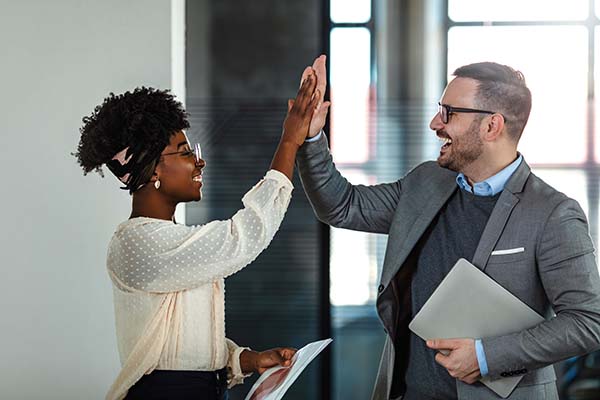 Two professionals giving a high-five in an office setting, one holding documents and the other carrying a laptop.